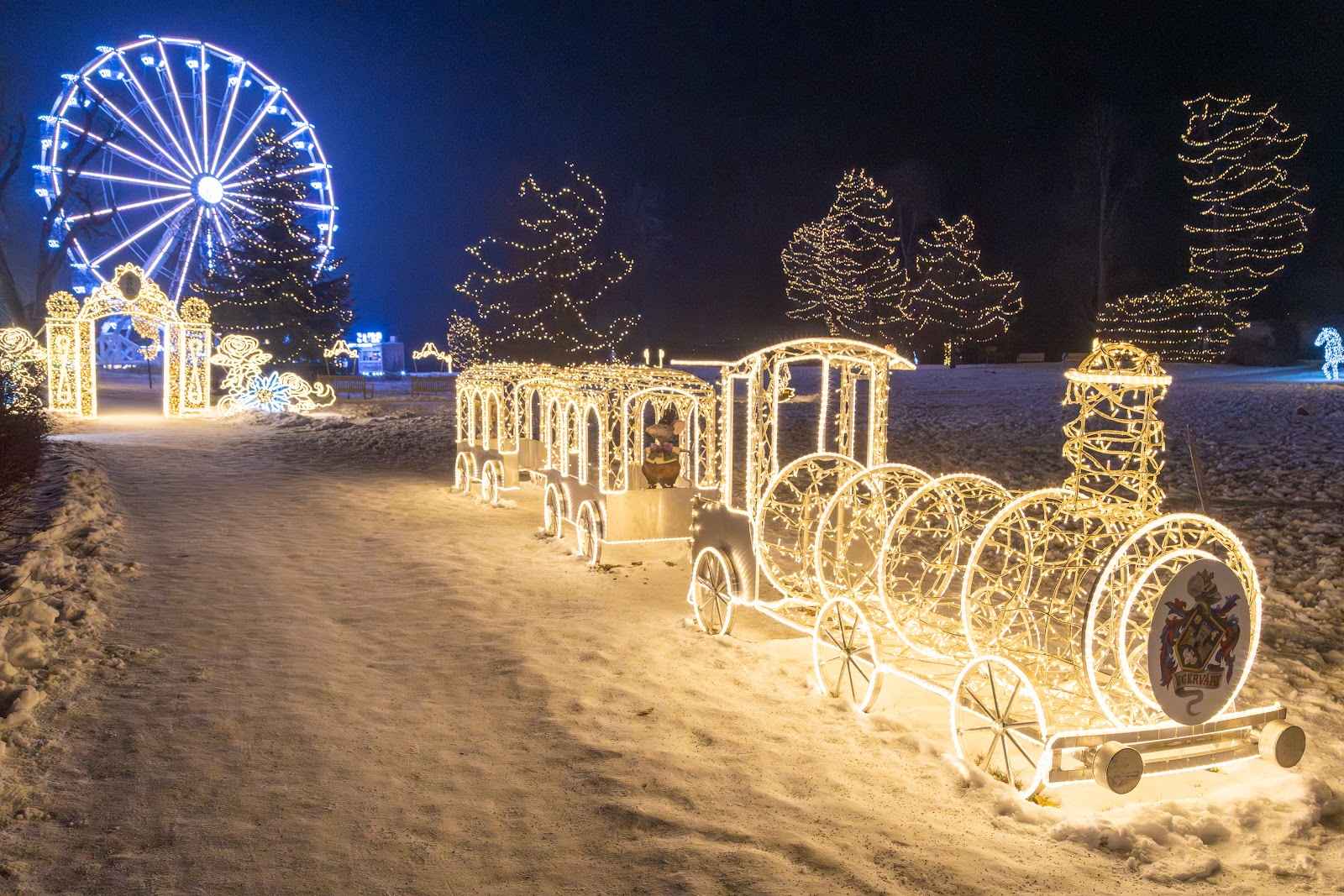 A locomotive and carriages made of light standing on a snow-covered path, a blue ferris wheel behind
