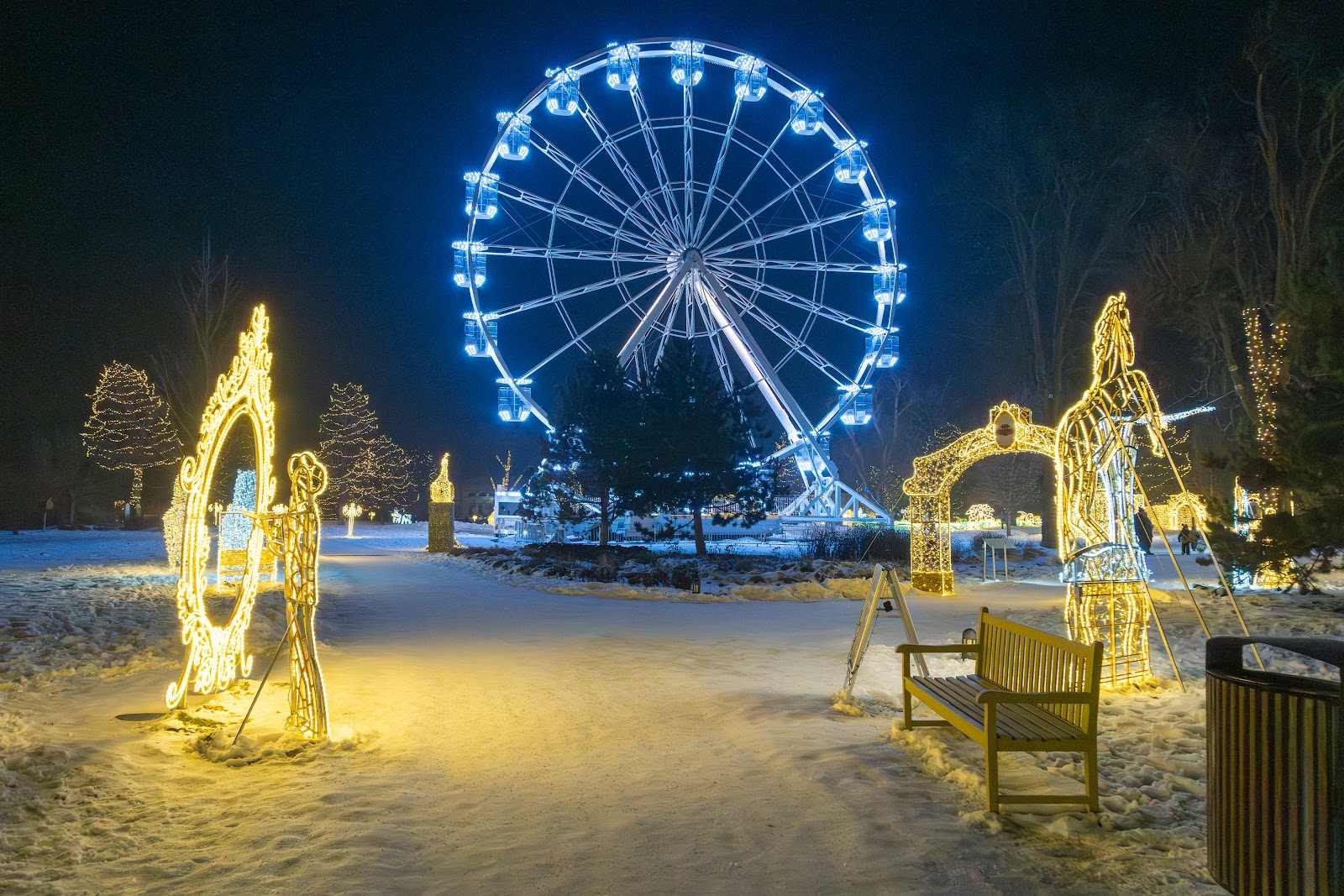 A lit ferris wheel and light sculptures scattered across a snow-covered palace garden